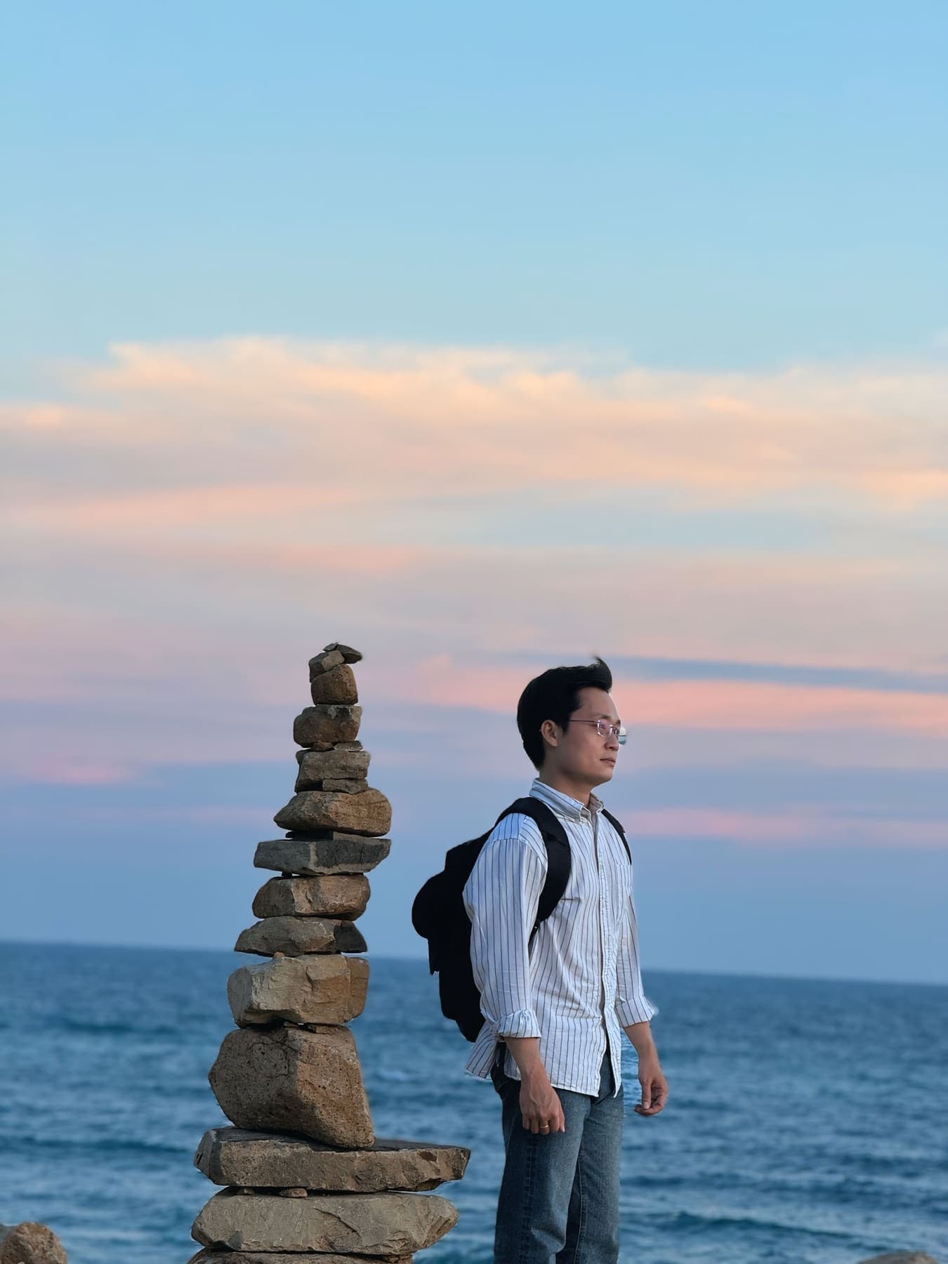 A man with a backpack beside a stack of stones by the sea at dusk