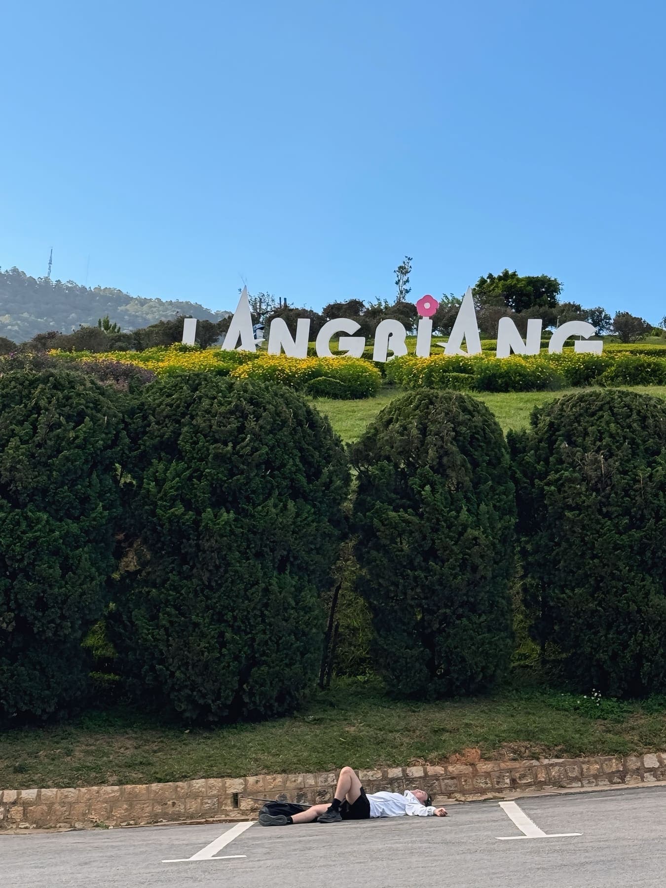 A hiker lying on the ground below a hillside sign reading “Langbiang”
