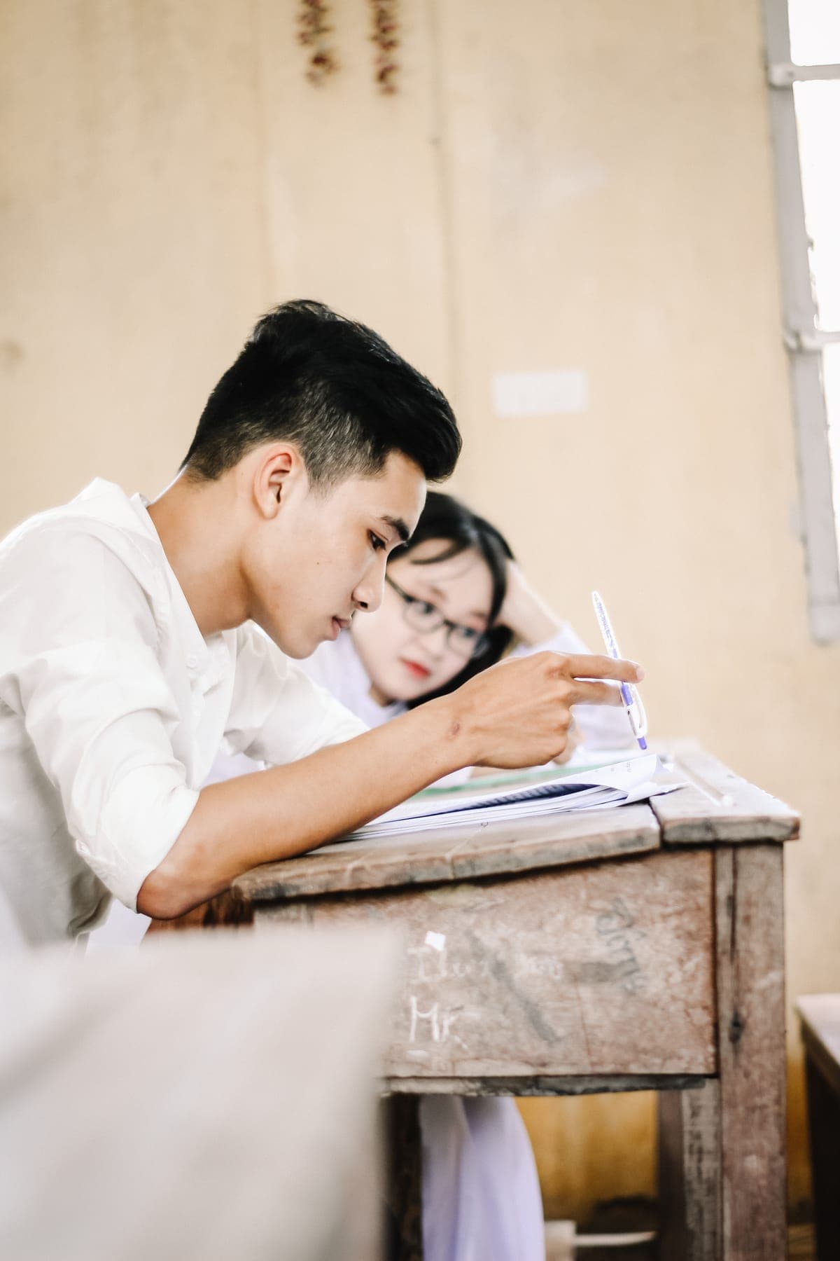 A boy writing at a wooden desk while a girl in glasses rests her head beside him