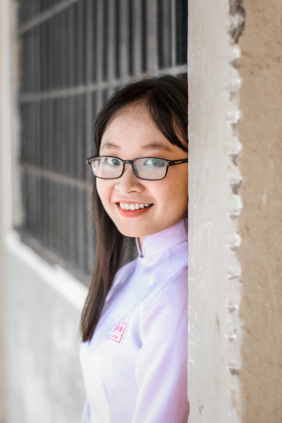 A smiling girl in glasses and an áo dài leaning against a pillar