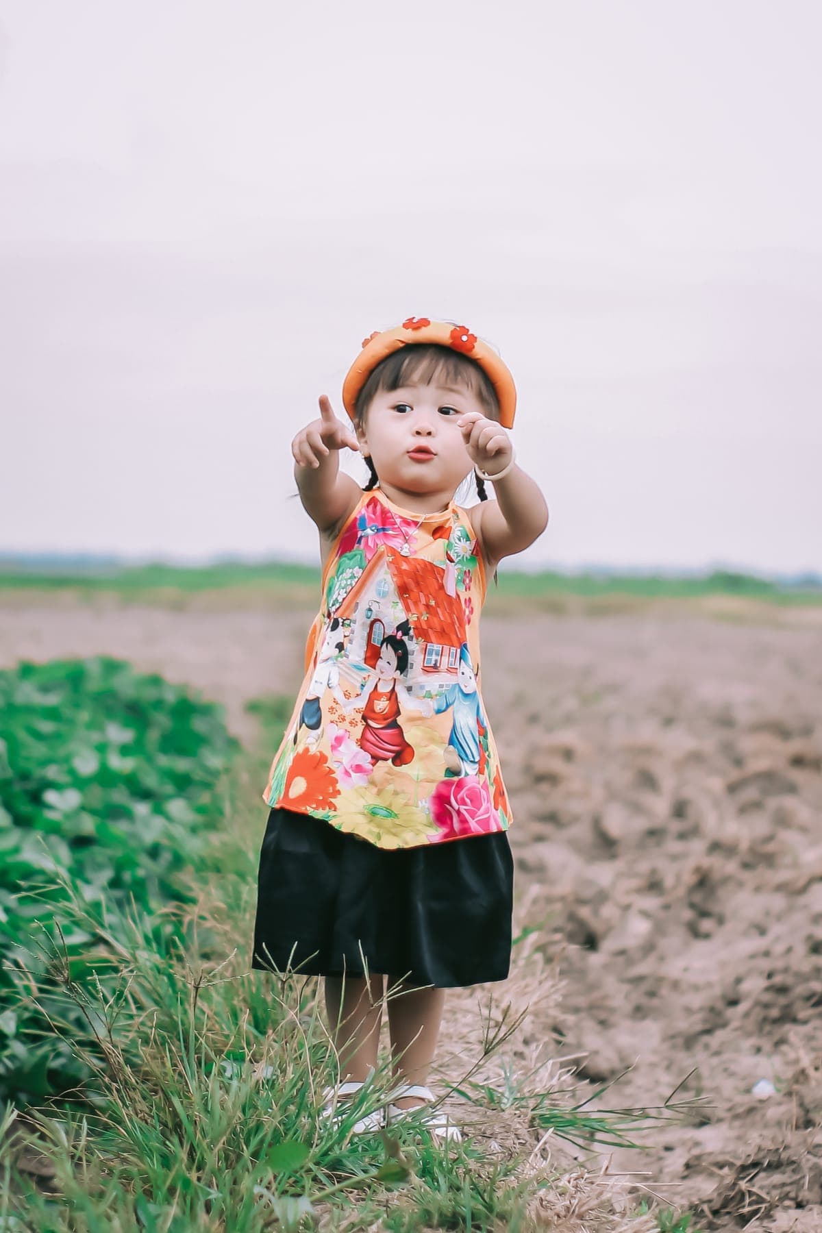 A toddler in a sun hat and floral top pointing at the camera in a field